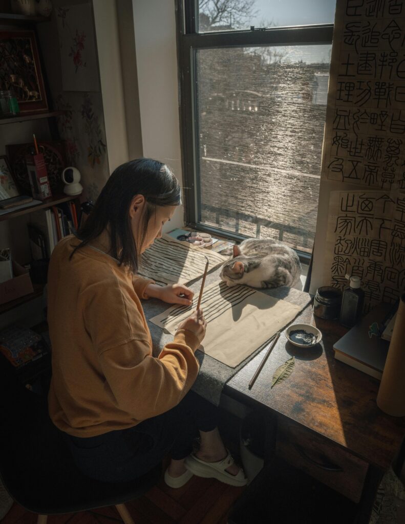 pexels photo 31069607 31069607 Asian woman practicing calligraphy by a sunny window, cat resting beside her. Cozy indoor scene.